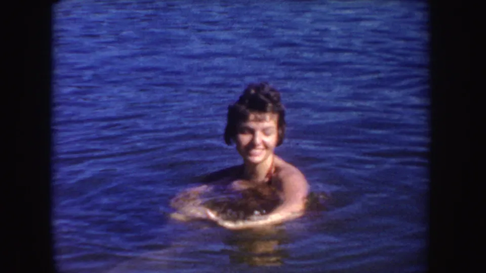 A smiling woman faces the camera while swimming in a South Dakota lake, shown in saturated 1954 8mm color.