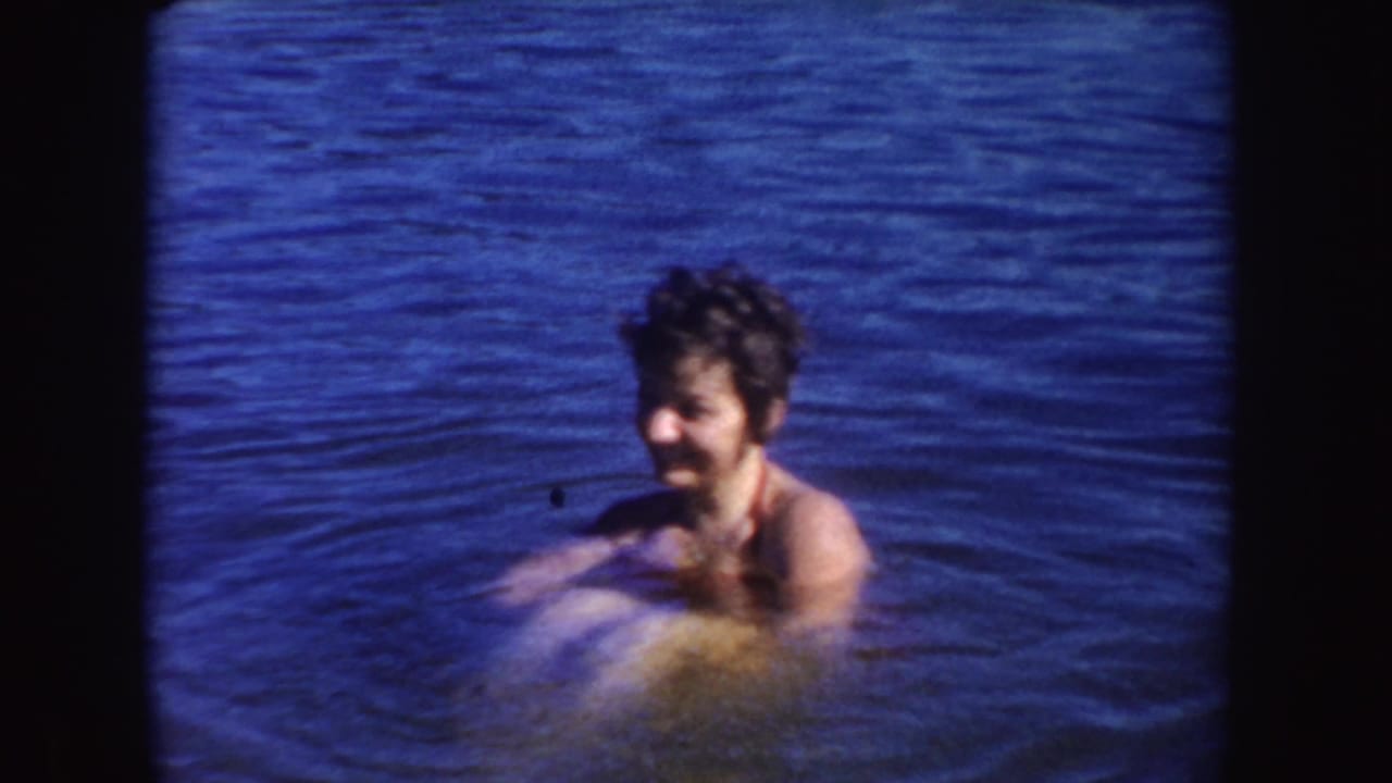 Woman in 1954 South Dakota lake footage turns slightly aside while smiling, with soft focus and saturated blue water.