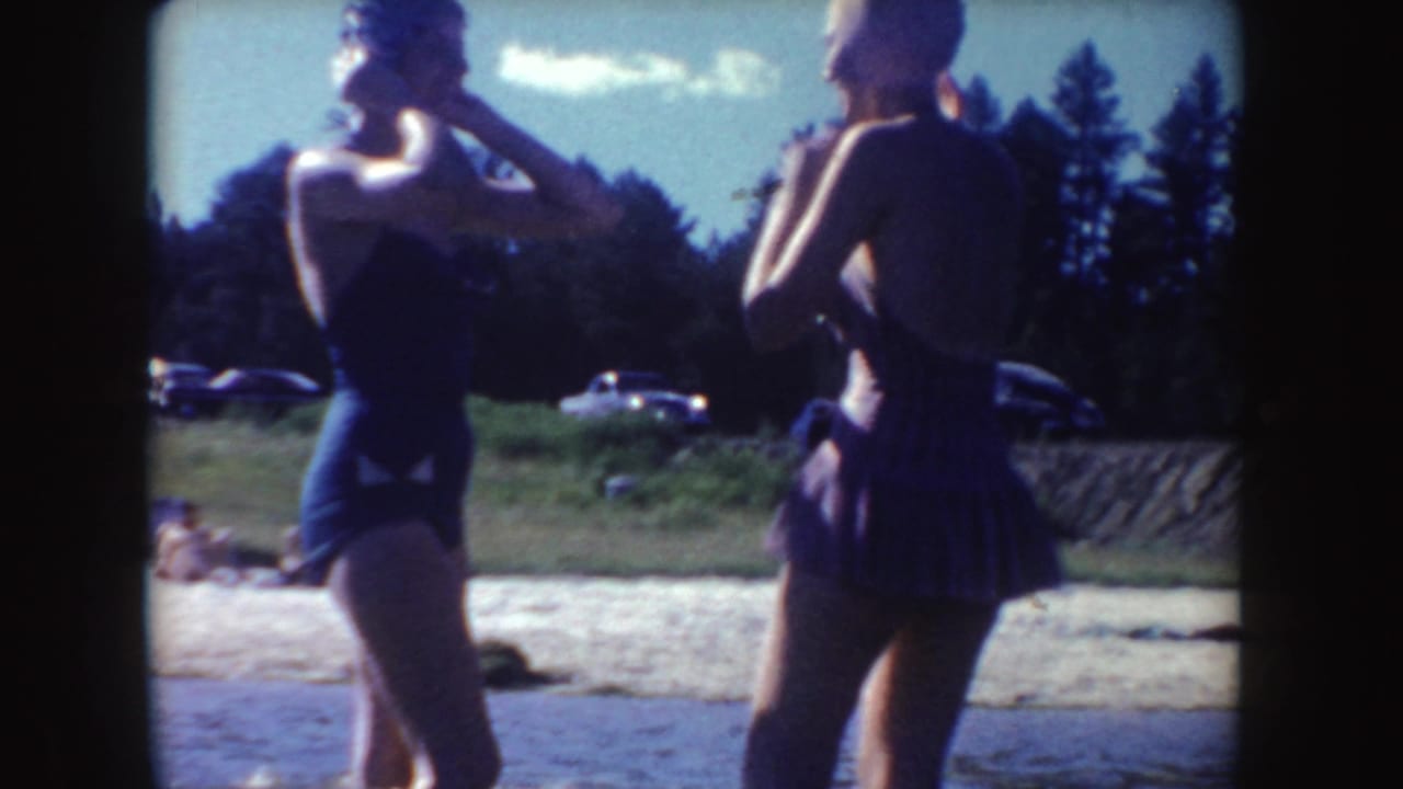 Two women in swimsuits stand near a sandy lakeshore with cars and trees behind them.