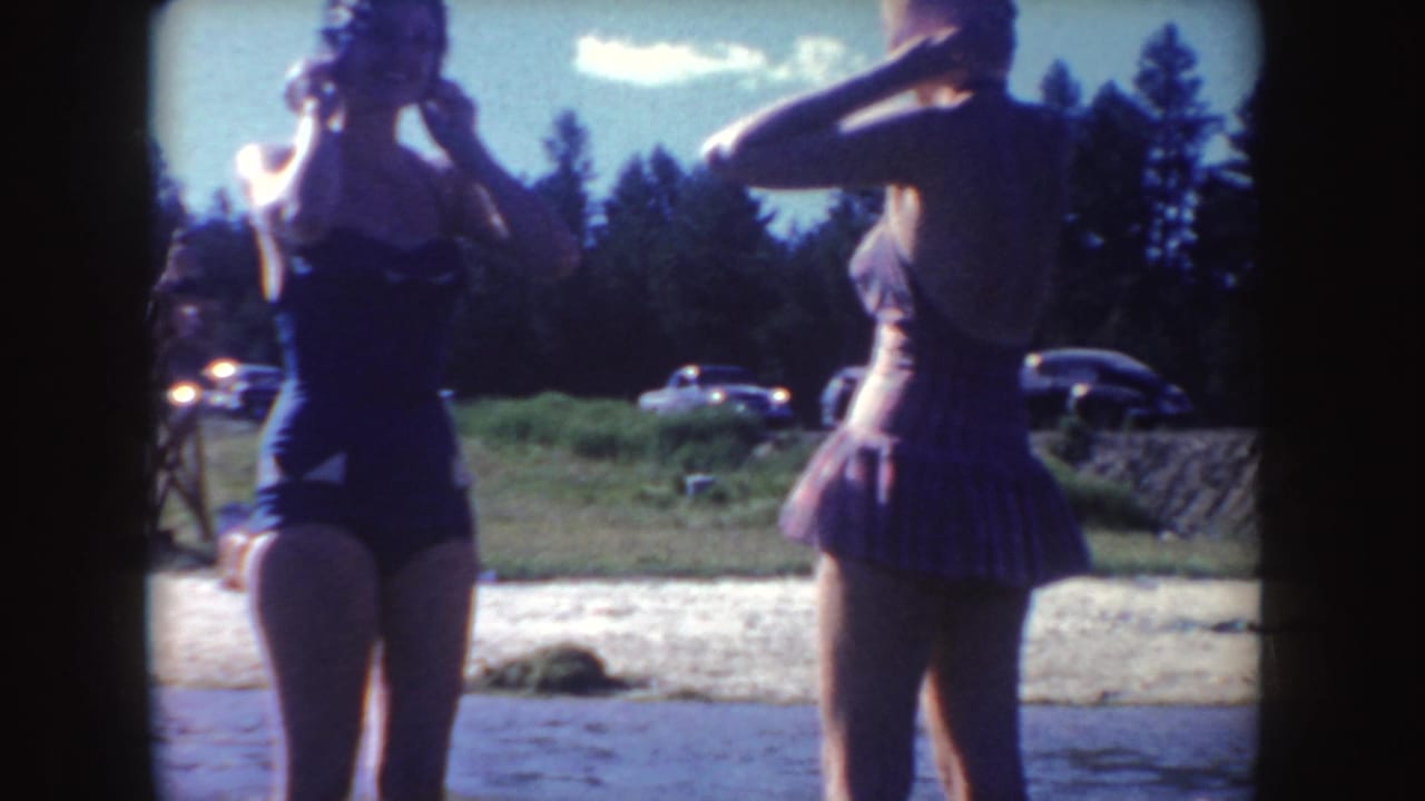 Two women in swimsuits stand at the edge of the water in the 1954 South Dakota home-movie scene, with vintage cars and trees behind them.