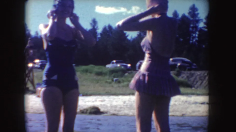 Two women in swimsuits stand at the edge of the water in the 1954 South Dakota home-movie scene, with vintage cars and trees behind them.
