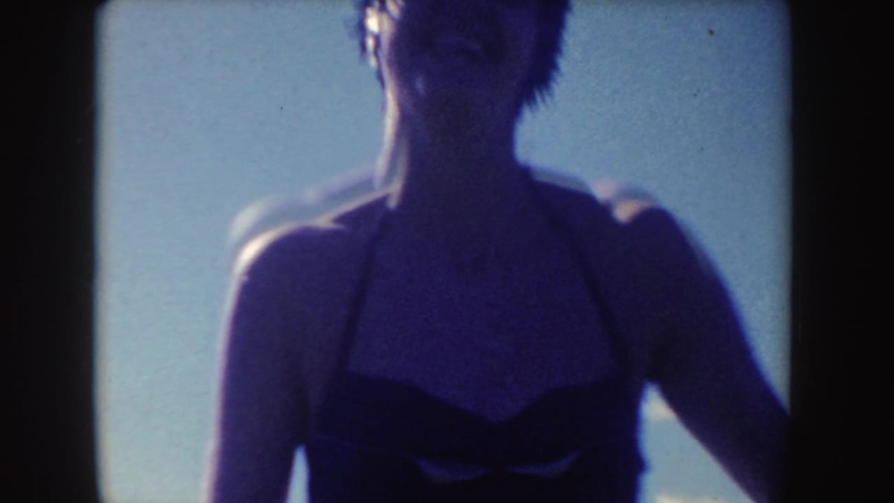 A close handheld 8mm view shows a woman in a dark swimsuit, softly focused against the bright lakeside sky.