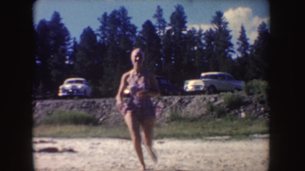 A woman in a patterned swimsuit walks across a sandy South Dakota lakeshore, with parked vintage cars and pine trees in the background.