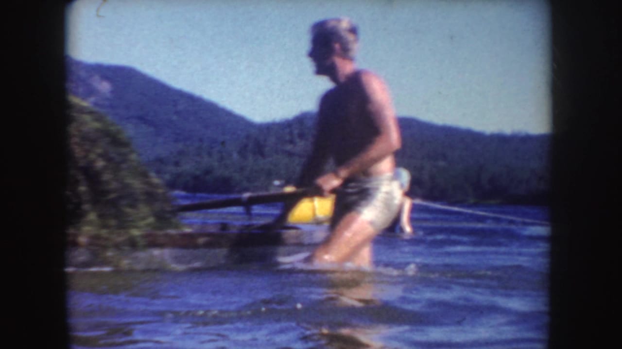 Adult man wading in a lake beside a yellow buoy with wooded hills behind him.