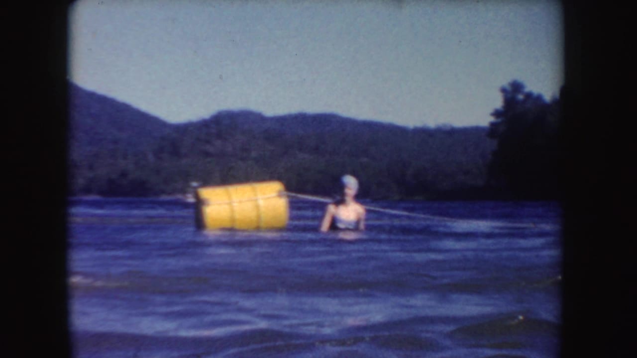In South Dakota 1954 home-movie footage, an adult woman swims near a yellow buoy in open lake water below wooded hills.