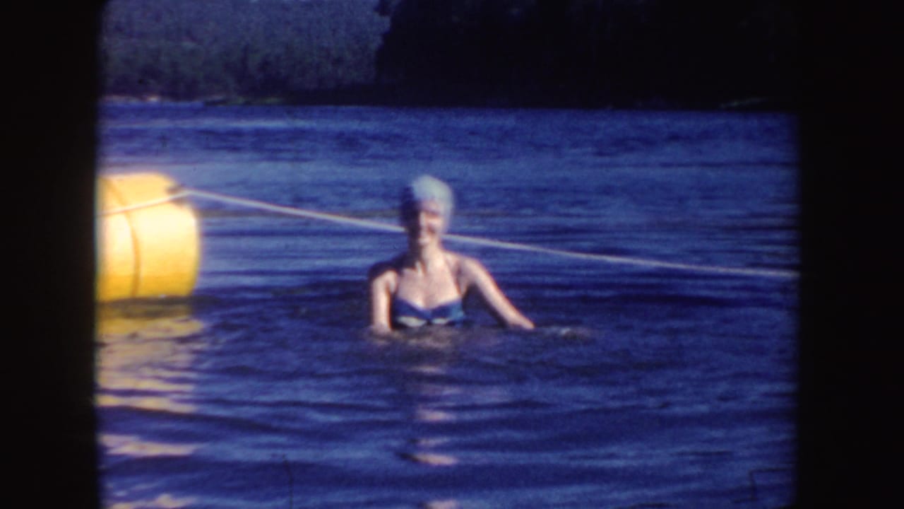 In South Dakota 1954 home-movie footage, an adult woman swims beside a buoy rope in lake water with wooded shoreline beyond.