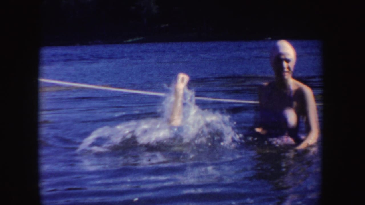 Two women in swim caps stand in lake water as one splashes or begins a handstand.