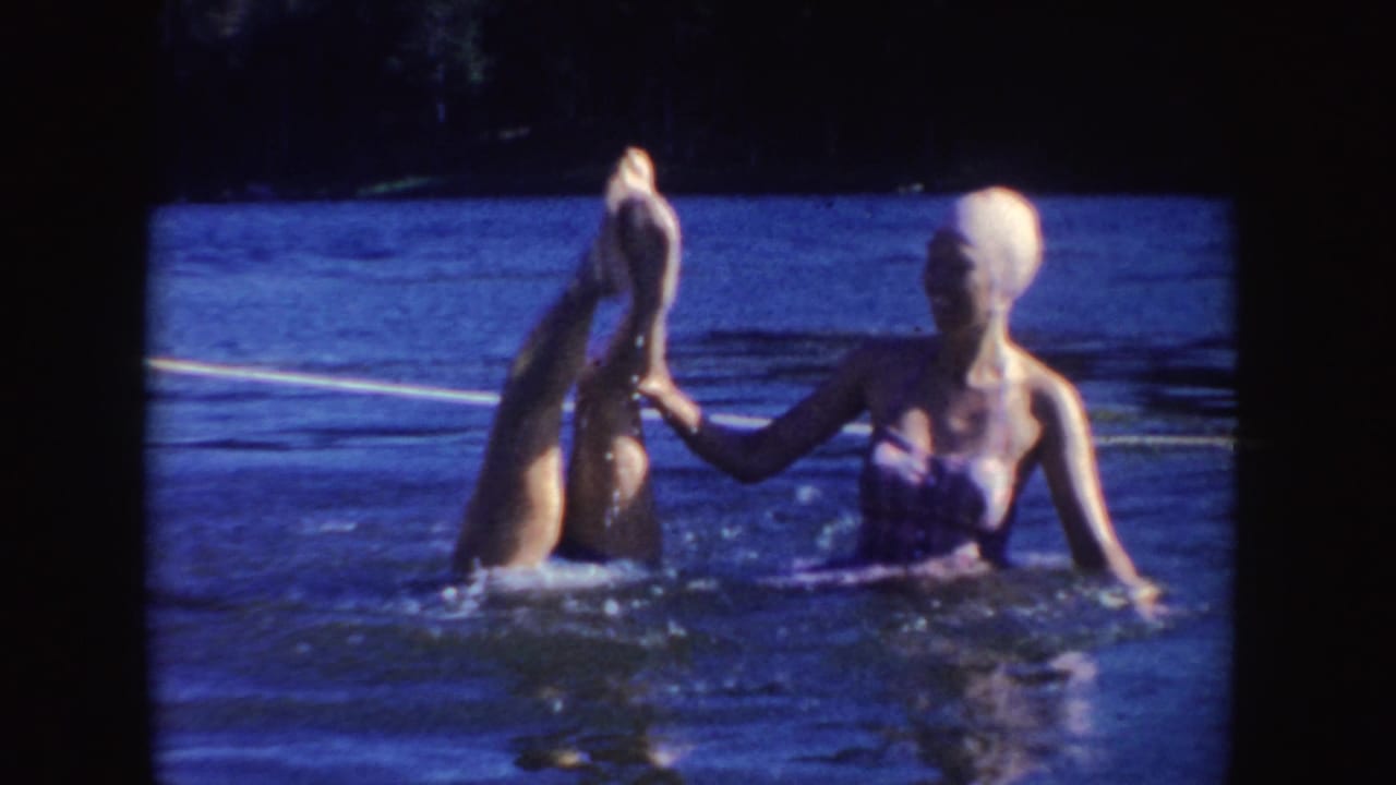 A woman performs a handstand in lake water while another woman steadies her leg.