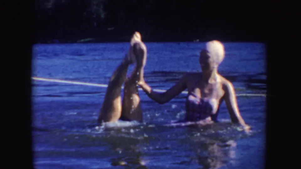 A woman in a swim cap performs a lake handstand while another woman in a bathing suit helps support her, with open water behind them.