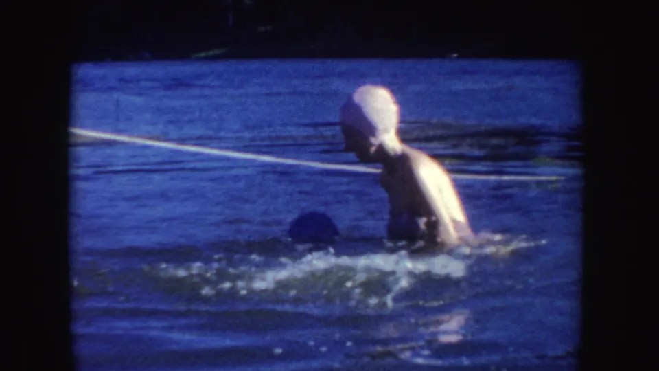 A woman in a white swim cap and bathing suit moves through the lake as another swimmer is partly submerged nearby.
