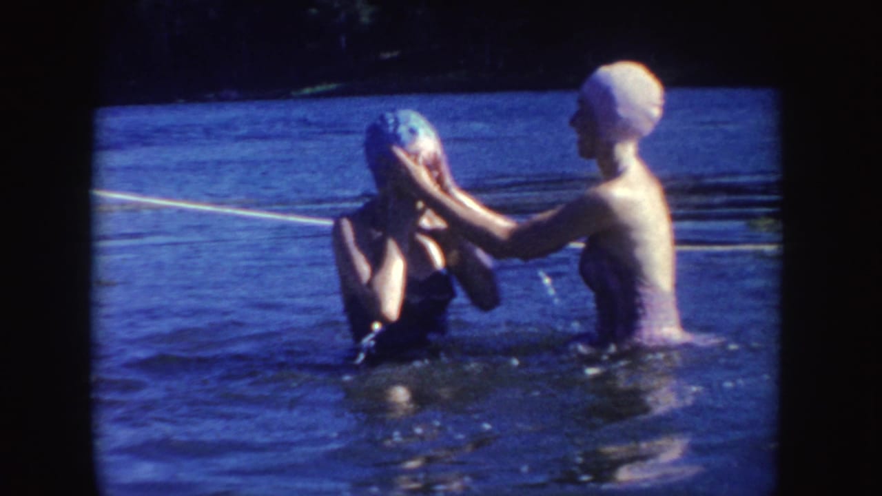 Two women in swim caps and bathing suits stand close together in a lake, with one appearing to adjust the other swimmer’s cap.