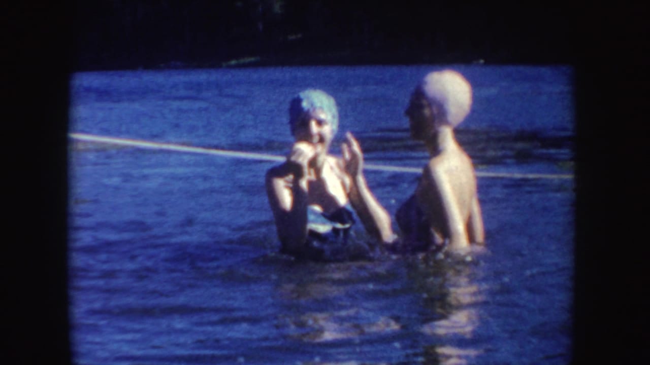Two women in swim caps stand chest-deep in lake water near a rope line.