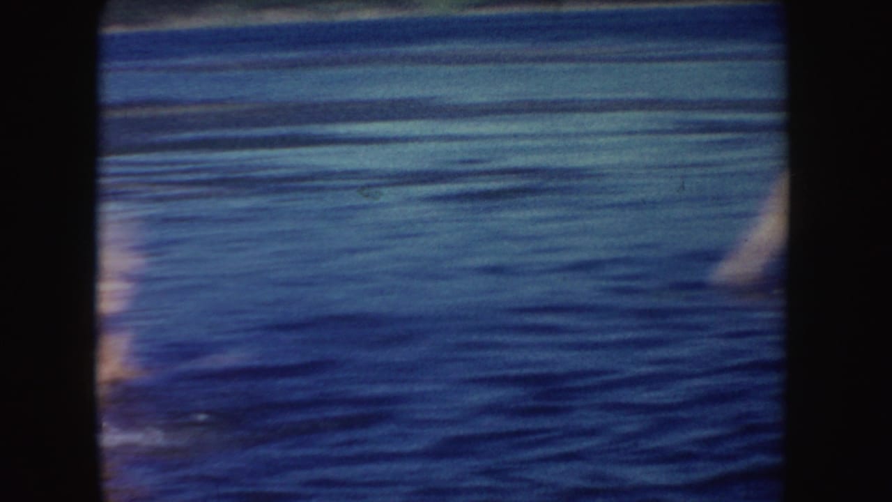 Handheld 8mm frame of rippling South Dakota lake water with partial blurred swimmers entering at the left and right edges.