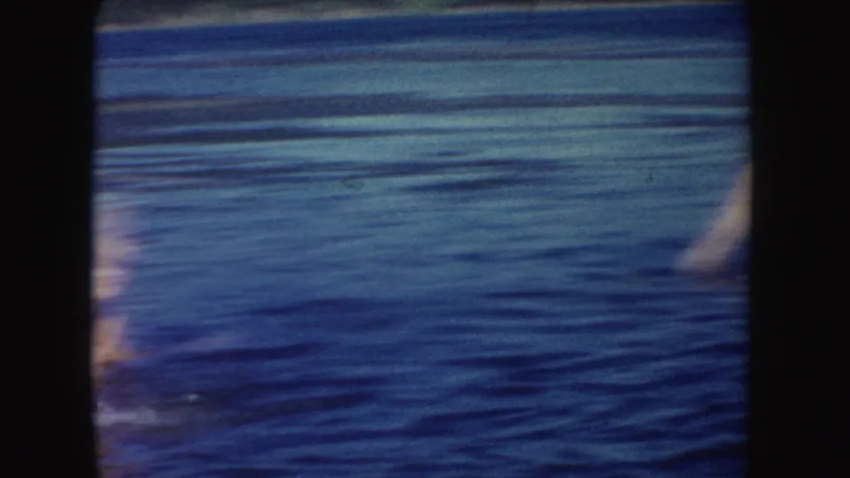 Handheld 8mm frame of rippling South Dakota lake water with partial blurred swimmers entering at the left and right edges.