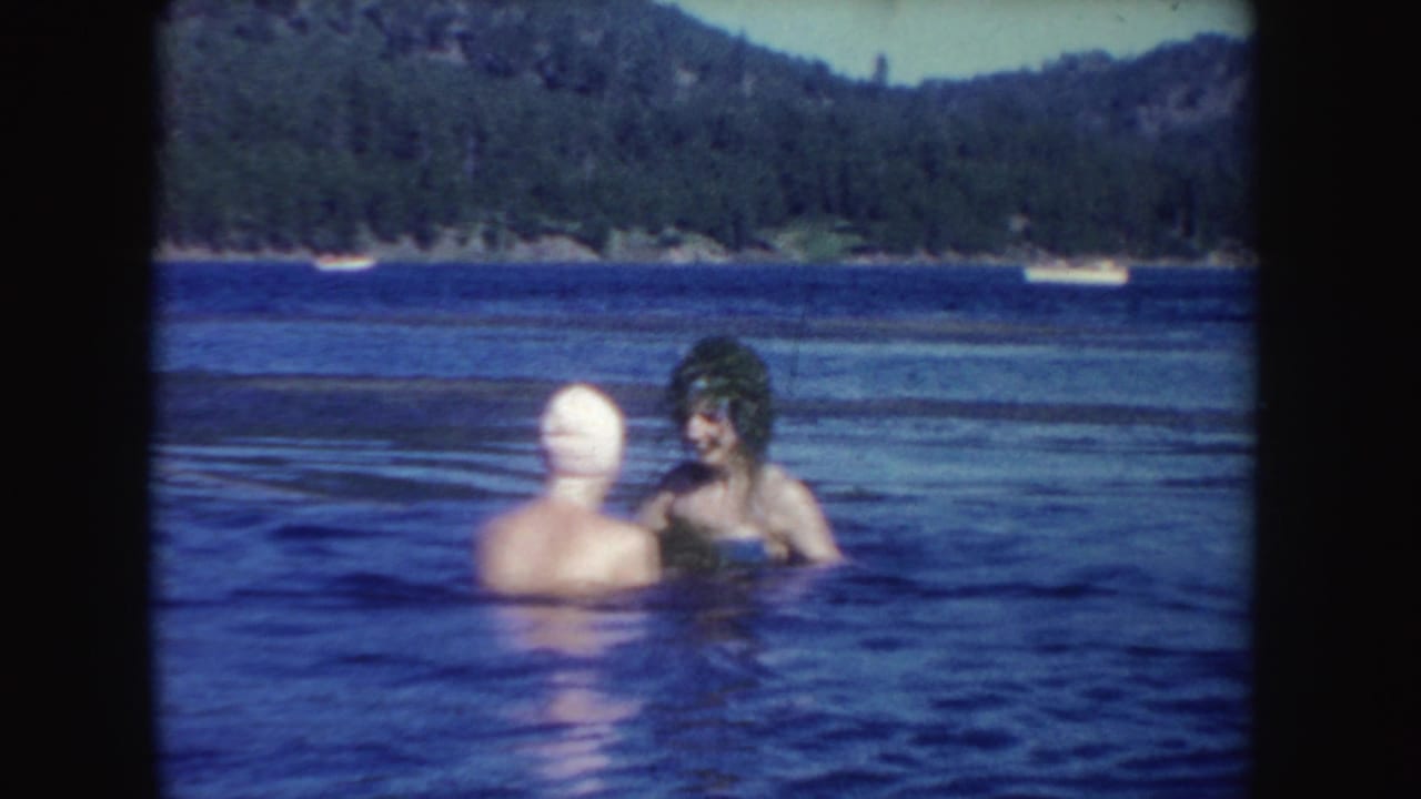 Two swimmers in a lake with wooded hills and small boats in the background.