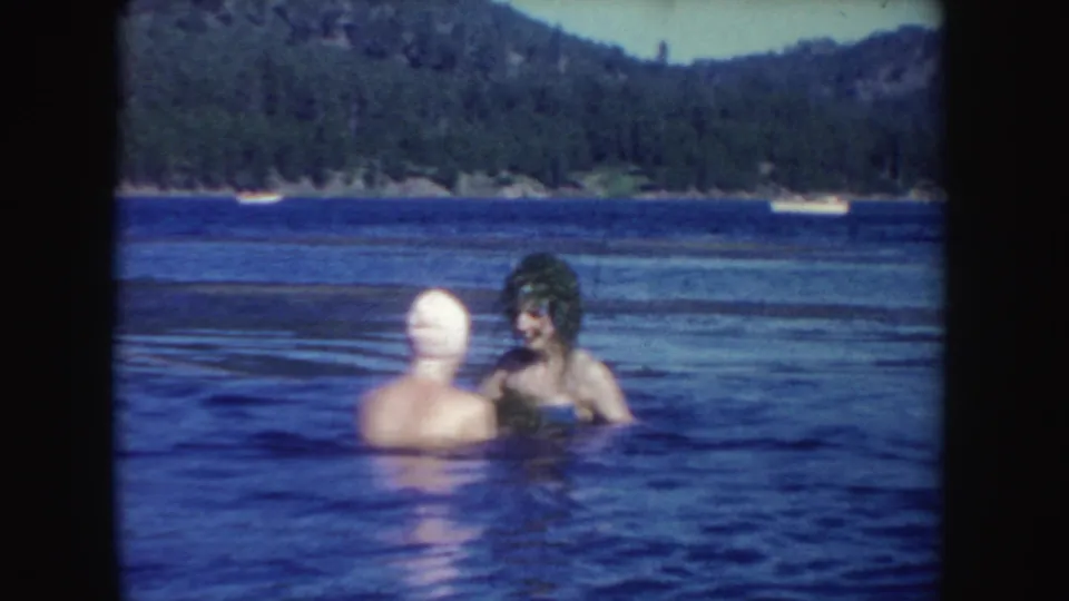 1954 South Dakota 8mm frame showing two young women chest-deep in lake water near a wooded shoreline.
