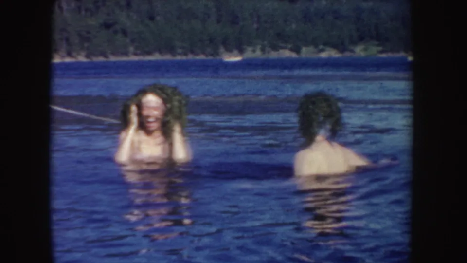 In 1954 South Dakota home-movie footage, two women are chest-deep in a lake as one laughs toward the camera with wet hair.