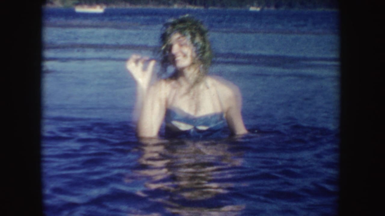 A woman in a blue swimsuit smiles in a South Dakota lake while holding up wet vegetation in the 1954 home-movie frame.