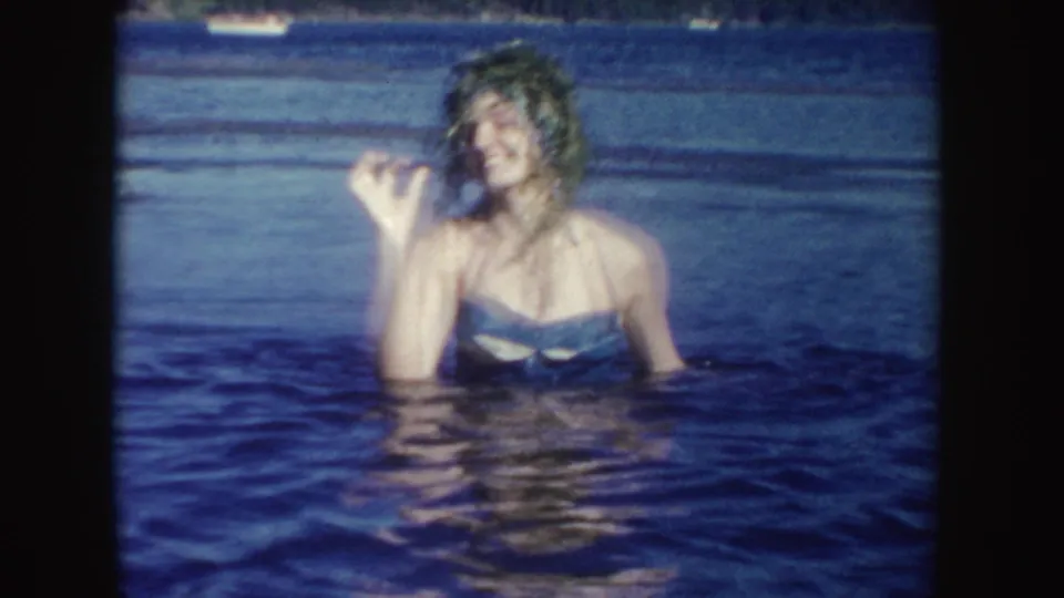 A woman in a blue swimsuit smiles in a South Dakota lake while holding up wet vegetation in the 1954 home-movie frame.