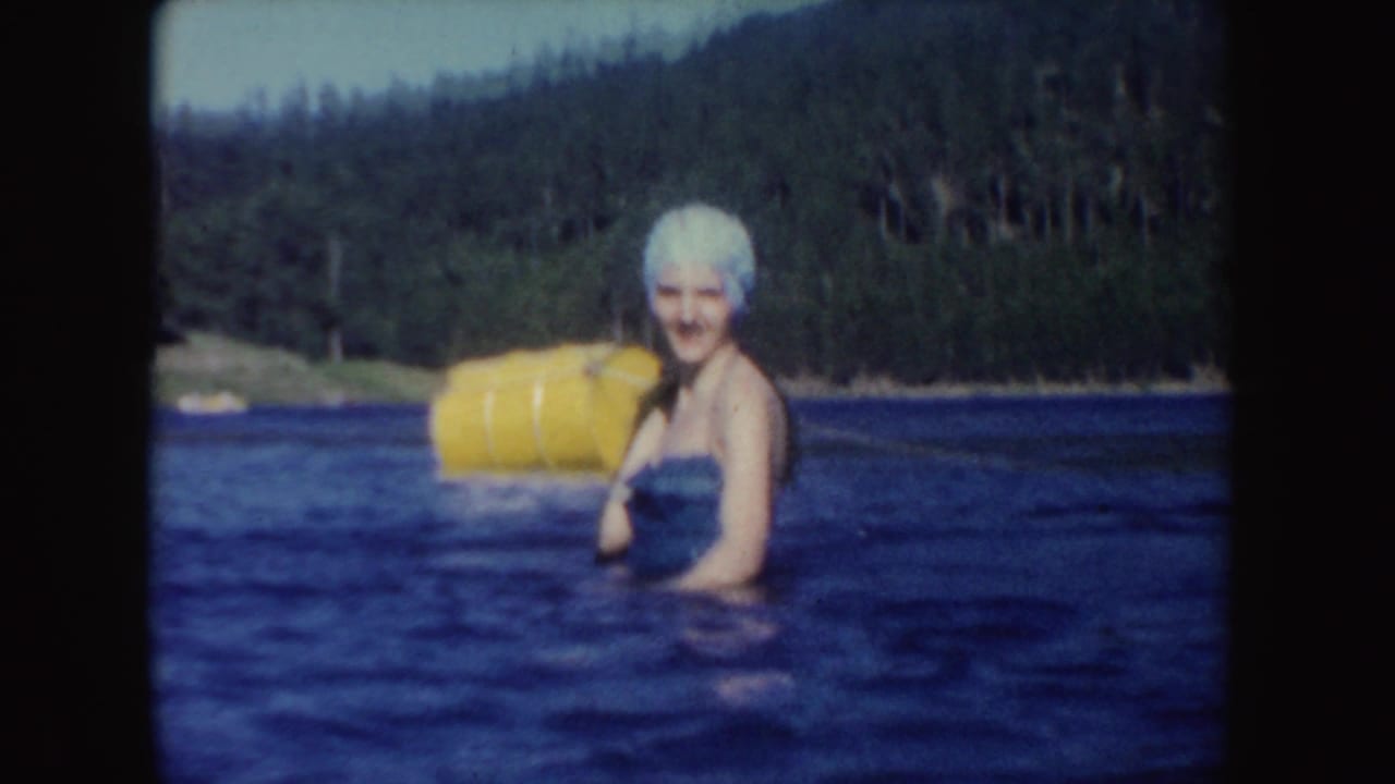A woman in a white swim cap and blue swimsuit stands in lake water near a yellow float, with trees and hills behind her.