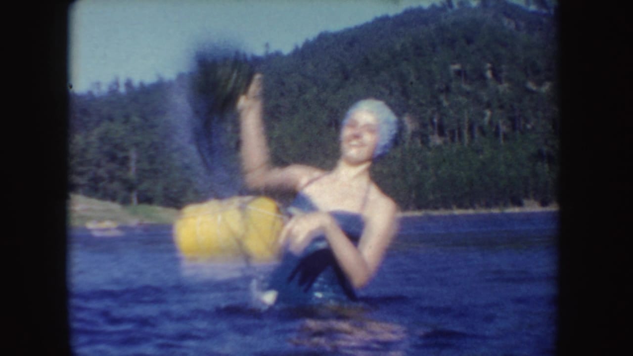 In the lake, a woman wearing a white swim cap laughs while lifting lake weeds and holding a yellow float against a wooded shoreline.