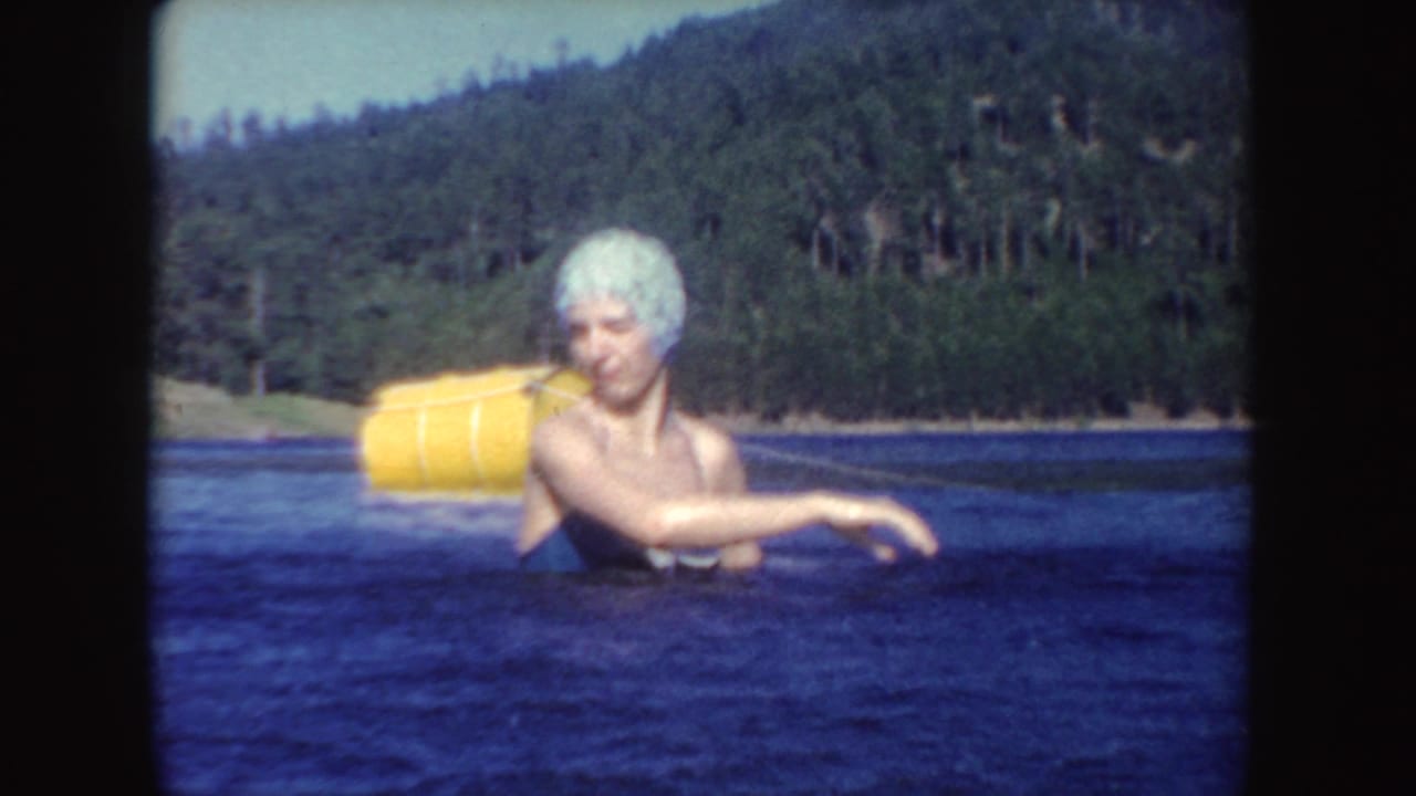 1954 South Dakota home-movie frame of a woman in a blue bathing suit and white swim cap wading near a yellow buoy.