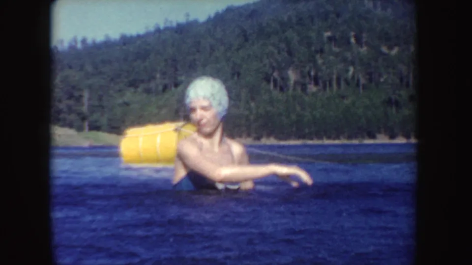 1954 South Dakota home-movie frame of a woman in a blue bathing suit and white swim cap wading near a yellow buoy.