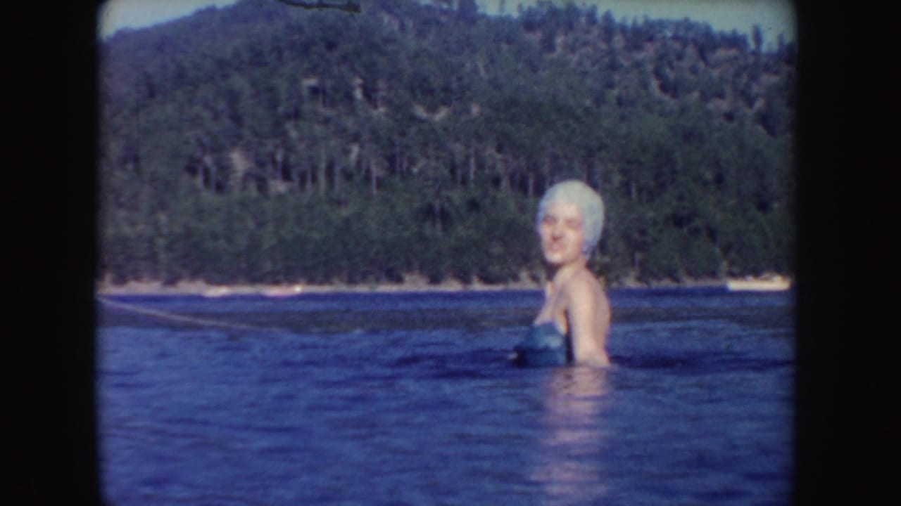 1954 South Dakota lake scene with a woman in a white swim cap standing chest-deep before a wooded hillside.