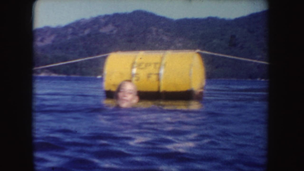 1954 South Dakota 8mm frame showing a swimmer near a large yellow buoy or depth marker on a wooded lake.