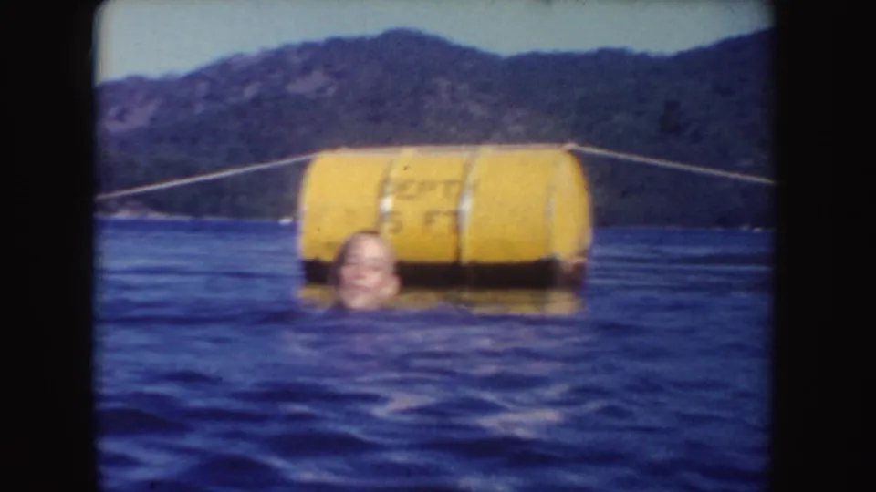 1954 South Dakota 8mm frame showing a swimmer near a large yellow buoy or depth marker on a wooded lake.
