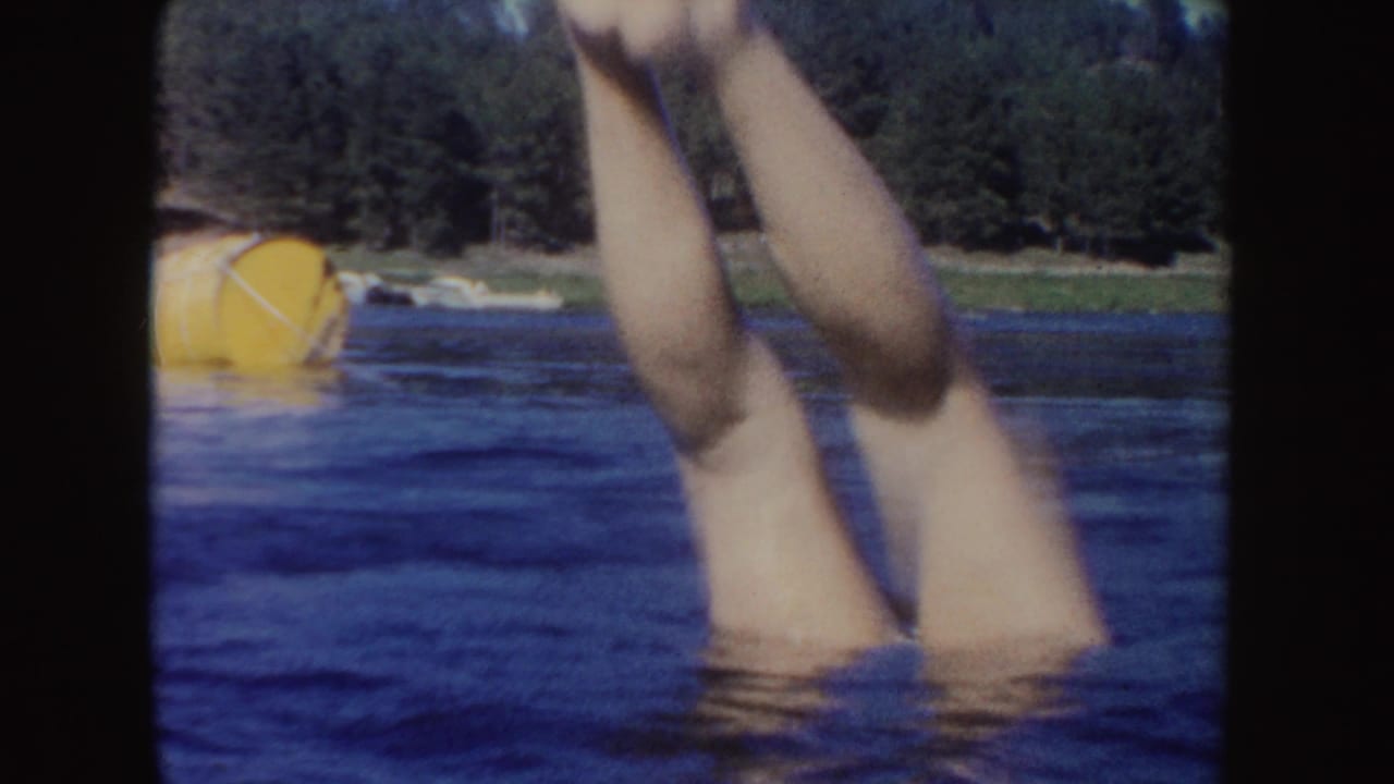 A swimmer's legs rise from the lake in a playful kick, with a yellow buoy and wooded shoreline visible behind.