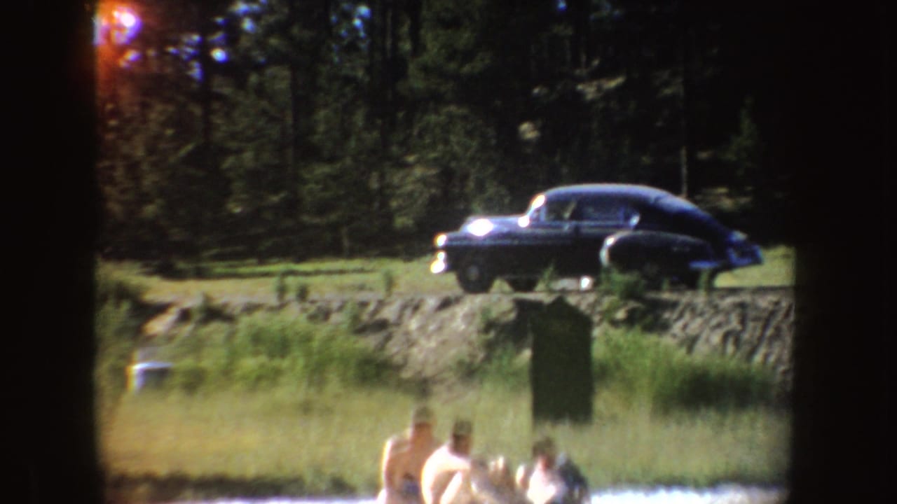 1954 South Dakota lakeside frame with a dark rounded sedan on a grassy rise above swimmers and a wooded background.