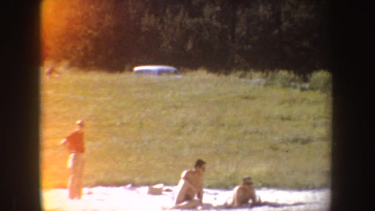 1954 South Dakota home-movie view of people relaxing near a sandy lake edge, with grass, trees, and warm film exposure.