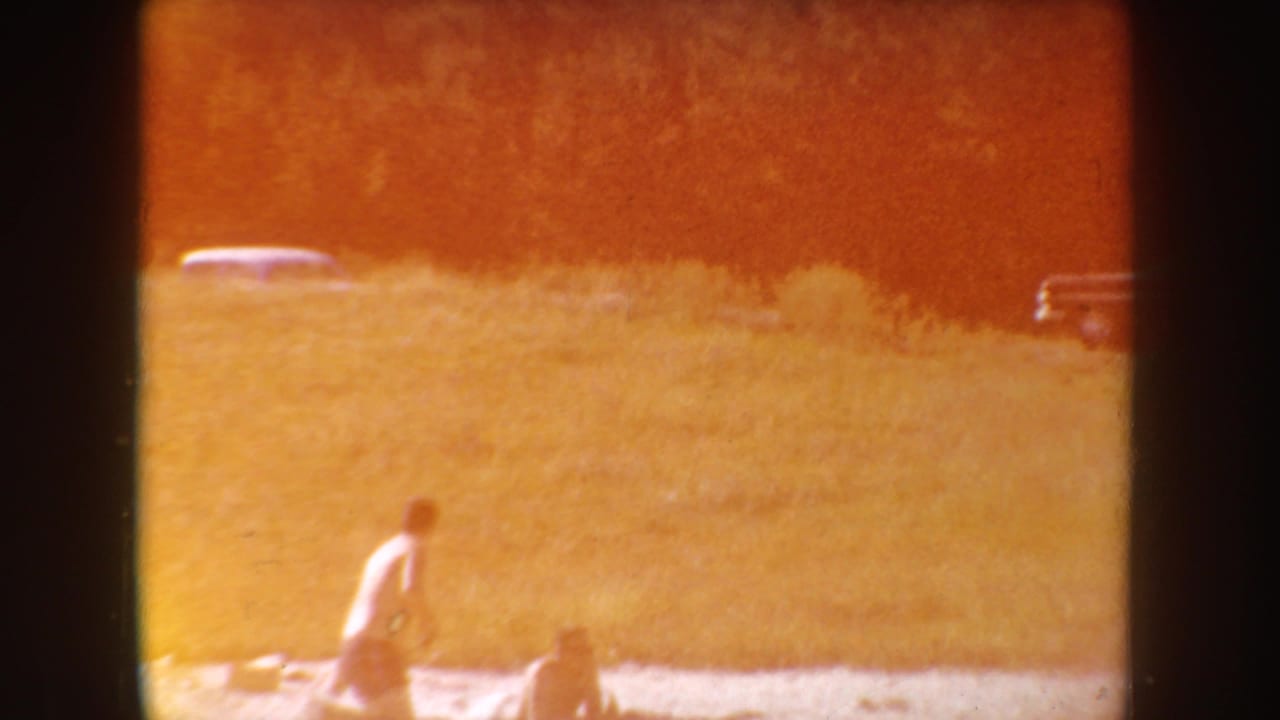 1954 South Dakota lakeside home-movie frame with strong warm flare, a person near the shore, grass, and distant parked cars.