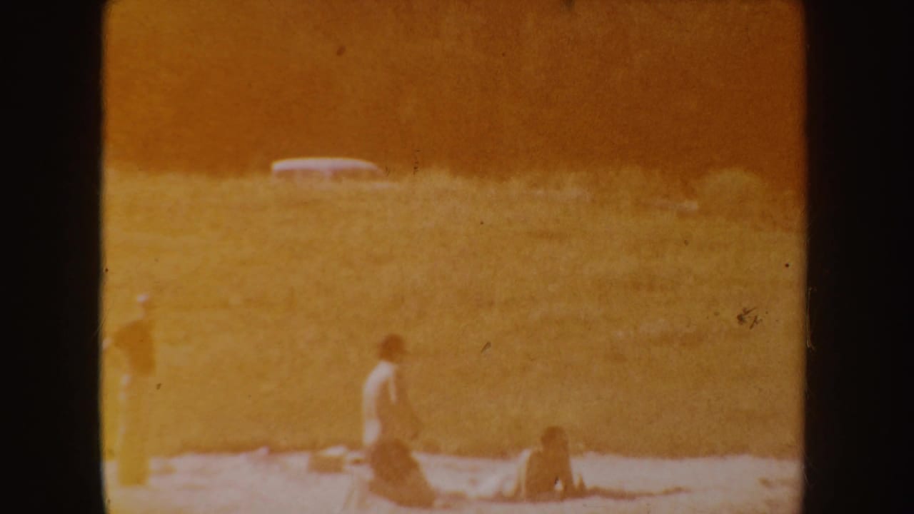 Faded lakeshore scene with figures on sand and a distant parked car.