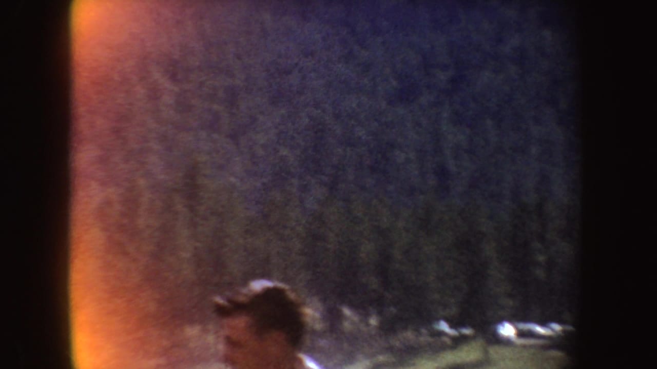 1954 South Dakota home-movie frame showing a partly cropped person in a grassy outdoor area with dark trees, hills, and orange film flare.