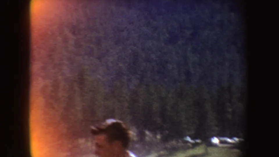 1954 South Dakota home-movie frame showing a partly cropped person in a grassy outdoor area with dark trees, hills, and orange film flare.