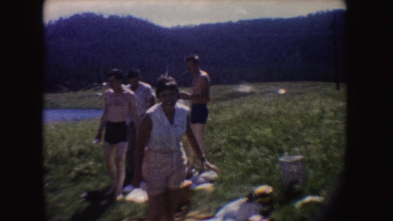 1954 South Dakota home-movie frame showing adults walking through a grassy area near water, with wooded hills and picnic items nearby.