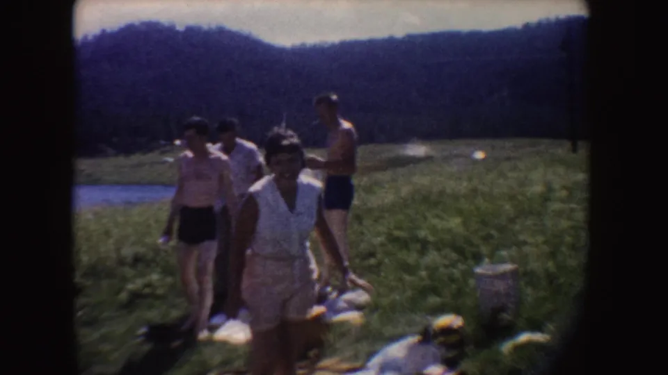 1954 South Dakota home-movie frame showing adults walking through a grassy area near water, with wooded hills and picnic items nearby.