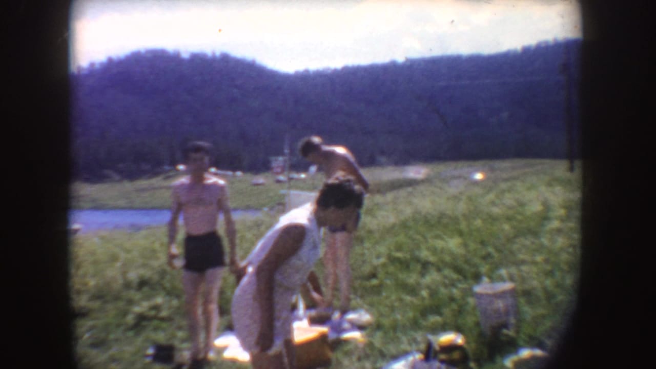 1954 South Dakota lakeside vacation frame with a woman bending toward belongings as two boys stand nearby on the grass.