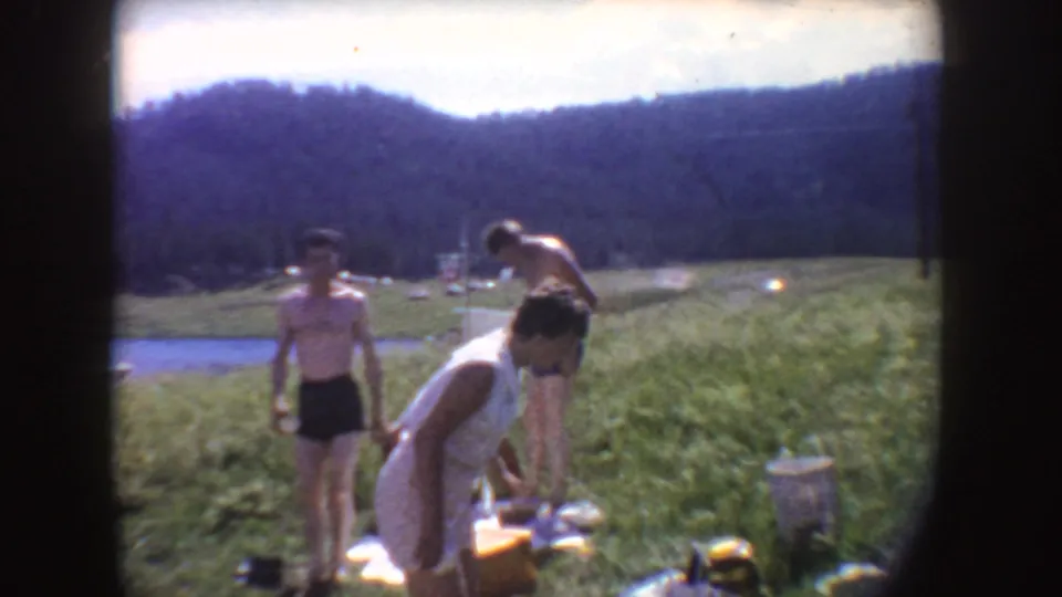 1954 South Dakota lakeside vacation frame with a woman bending toward belongings as two boys stand nearby on the grass.