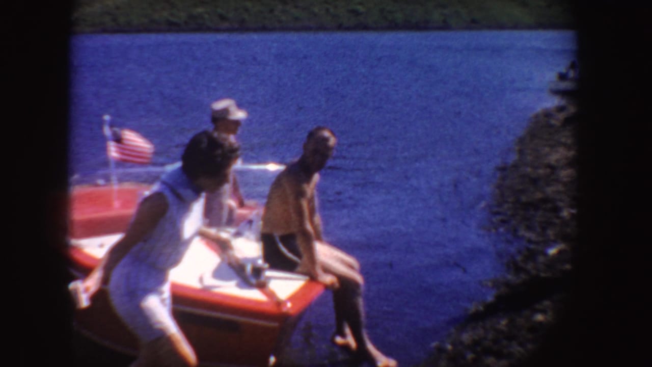 1954 South Dakota frame showing a woman in white beside a red motorboat on blue lake water, with seated passengers and a small American flag.