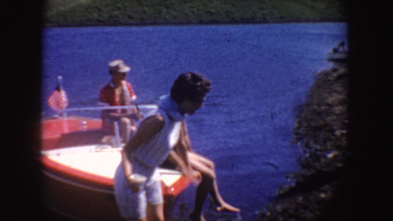 1954 South Dakota home-movie frame with a woman leaning near a red motorboat, seated passengers, blue lake water, and a small American flag.