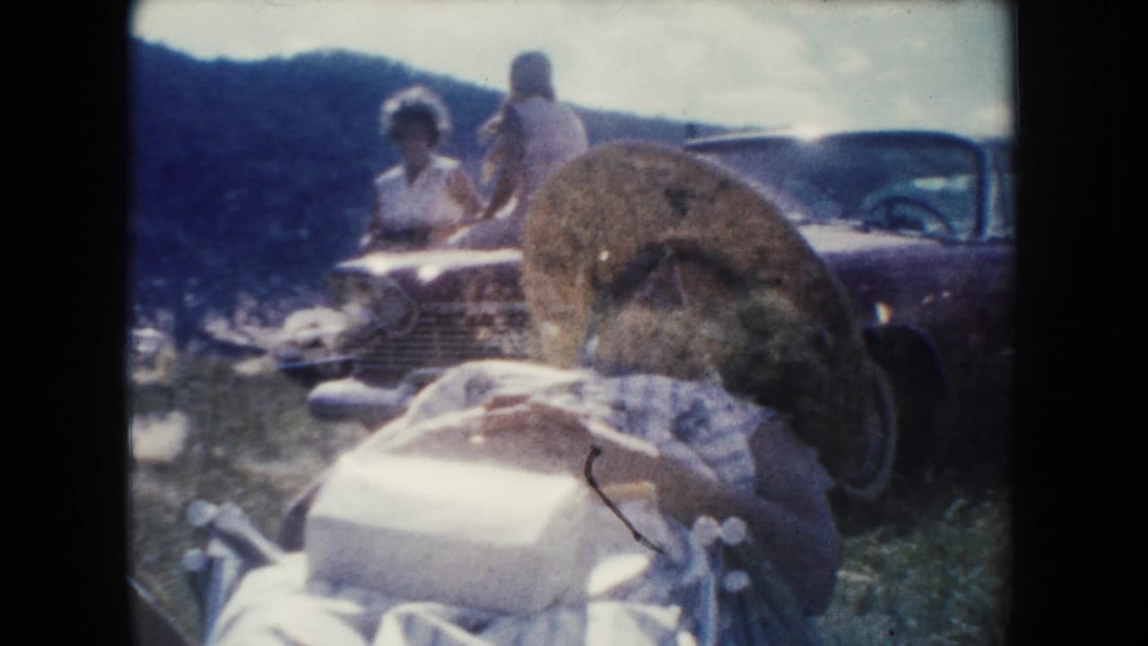 South Dakota home-movie frame with a woman lying on a blanket in front of a vintage car while two people stand or sit nearby.