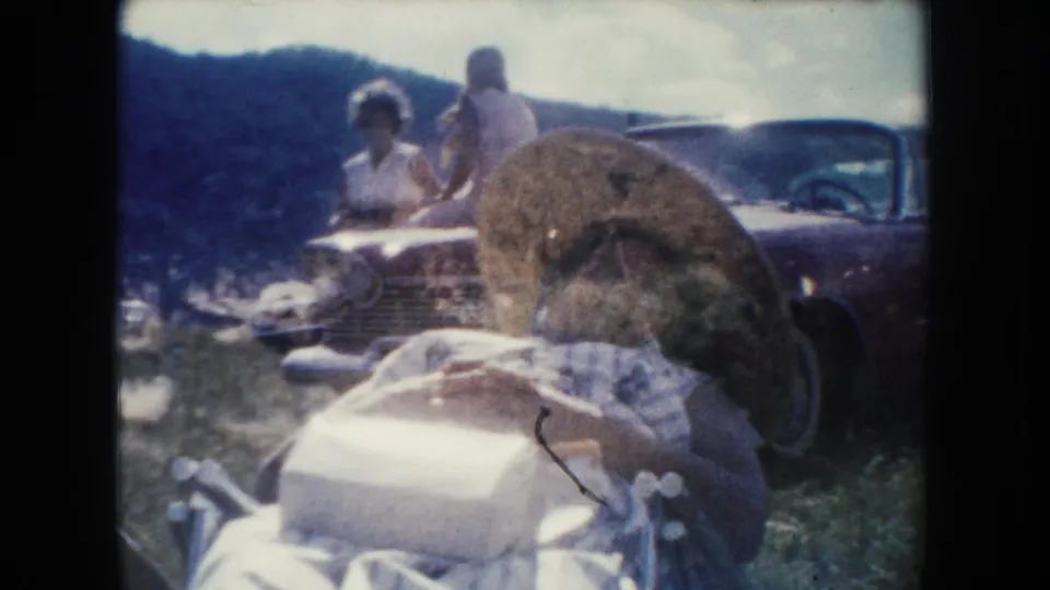 South Dakota home-movie frame with a woman lying on a blanket in front of a vintage car while two people stand or sit nearby.