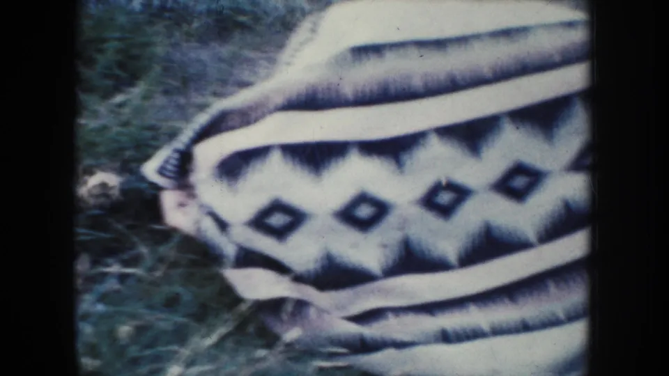 Close home-movie detail of a striped and diamond-patterned textile lying on grass, likely part of the outdoor resting setup.