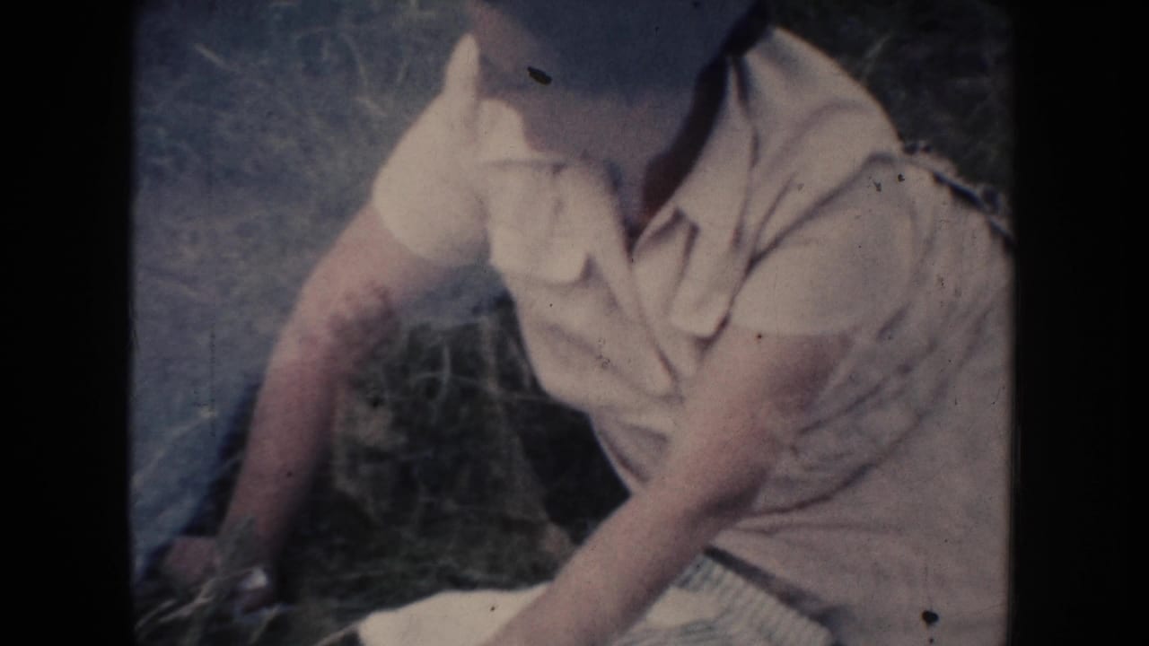 In 1954 South Dakota footage, a woman in a white dress sits on grass while handling a checked cloth.