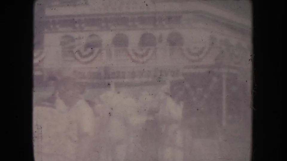 Faded 1954 South Dakota frame with several people standing near a bunting-decorated building in bright daylight.