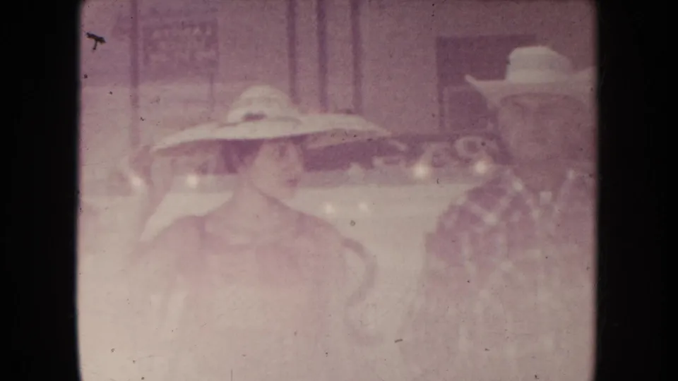 Faded 1954 South Dakota frame showing the same two adults in hats, with storefront details softened by exposure.
