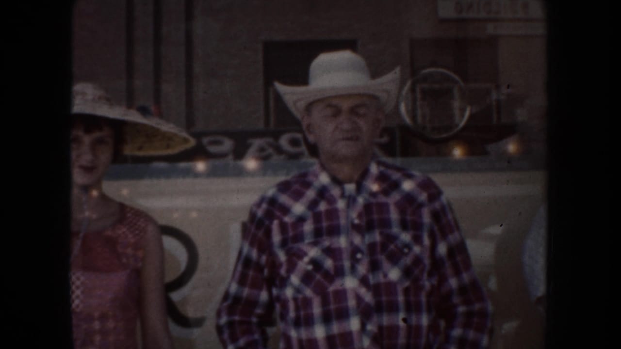 Man in a white cowboy hat and plaid shirt stands beside a partially visible woman near a storefront.